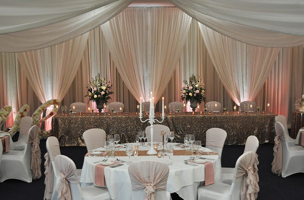 2 layer drapes to frame wedding flowers for a pink and gold uplit wedding breakfast at the De Vere Cotswold Water Park with ceiling drapes and tied back wall drapes