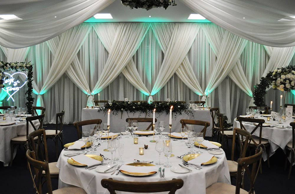 Cross over wall drapes with foliage and green uplighting, neon sign and archway at a wedding at De Vere Cotswold Water park Hotel