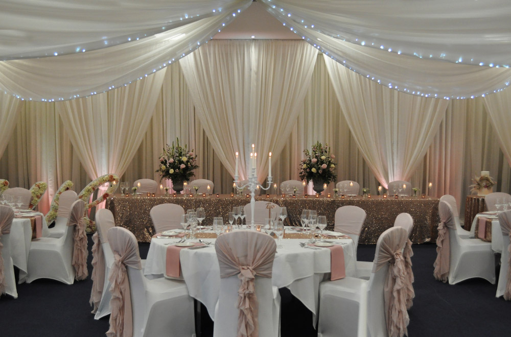 Wall and Ceiling drapes for a wedding with pink ruffle hoods and gold sequin table at De Vere Cotswold Water Park hotel with fairy lights and LEd uplighters